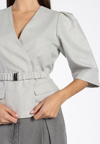 Lichtgrijze, gestructureerde wikkeltop met een V-hals, pofmouwen en een geplooide riem met een zilveren gesp. Twee voorzakken zijn zichtbaar.