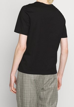 Person trägt ein einfaches schwarzes Kurzarmshirt und graue karierte Hosen, steht mit dem Rücken zur Kamera vor einem hellen Hintergrund.