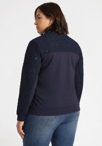 Jachetă bleumarin cu țesătură texturată și strălucitoare pe umeri și mâneci, cu un corp neted și manșete cu nervuri. Asortată cu blugi.