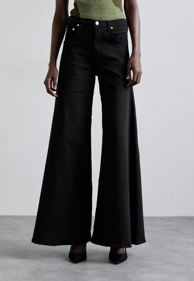 Pantaloni din denim negri cu croi larg și talie înaltă, cu buzunare frontale și accesorii din metal argintiu, asortați cu tocuri negre ascuțite.