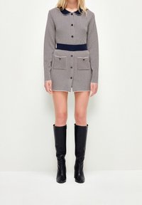 Abito a quadri con colletto, chiusura frontale con bottoni e due tasche frontali. Patta in blu navy. Modello che indossa stivali neri in pelle al ginocchio.