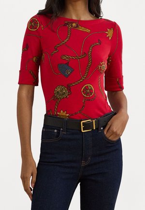 Top ajustado rojo con mangas cortas que presenta un estampado gráfico en dorado y azul. Combinado con vaqueros azul oscuro y un cinturón negro con hebilla dorada.