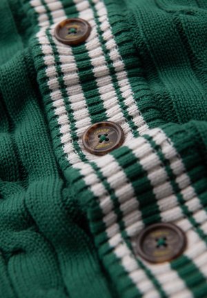 Tessuto lavorato a maglia verde con strisce bianche. Quattro bottoni marroni con un design a croce e testo in rilievo vicino al centro. Pattern testurizzato visibile.