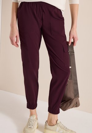 Personne portant un pantalon cargo violet foncé et des baskets beige, tenant un sac bandoulière taupe par sa sangle.