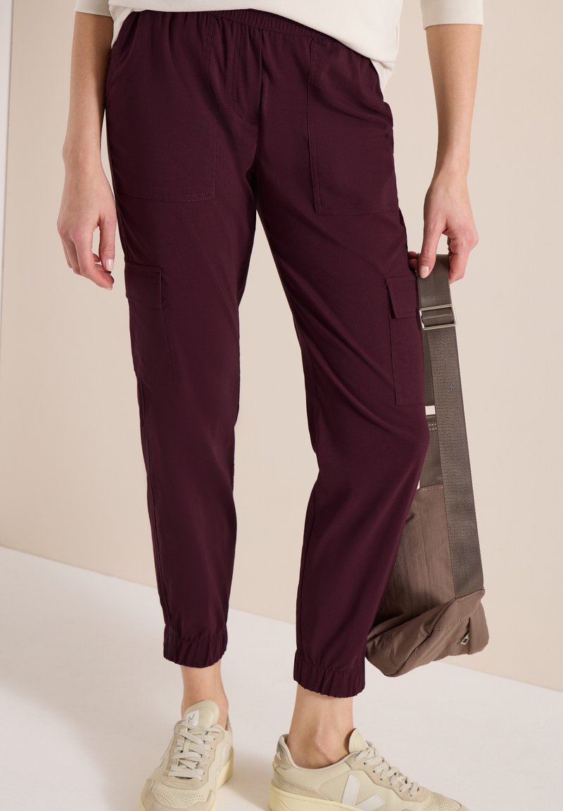 Personne portant un pantalon cargo violet foncé et des baskets beige, tenant un sac bandoulière taupe par sa sangle.