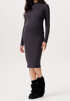 Donna che indossa un vestito midi grigio scuro aderente a maniche lunghe e stivaletti neri alla caviglia con lacci, su uno sfondo chiaro e semplice.