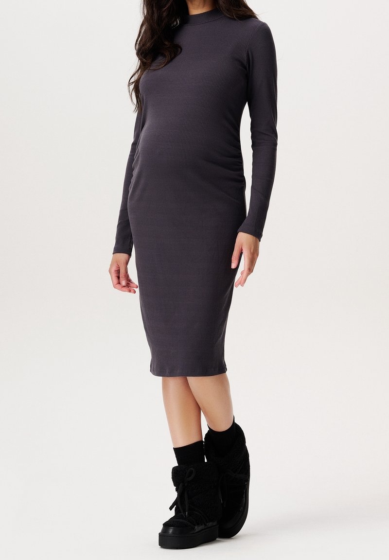 Femme portant une robe midi gris foncé à manches longues et ajustée, ainsi que des bottines noires montantes à lacets, sur un fond clair uni.