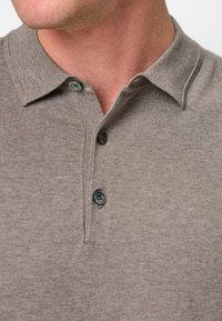 Ljust brunt polo-shirt med krage, tre svarta knappar och gröna detaljer. Tyget har en ribbad textur och en mjuk yta.