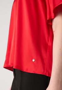 Top rosso a maniche corte realizzato in tessuto liscio, con orlo arrotondato, dotato di un piccolo logo argentato all'orlo inferiore. Pantaloni neri parzialmente visibili.