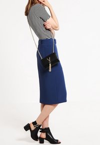 Randig kortärmad topp, blå knä-lång kjol, svart axelväska med fransar och svarta klacksandaler med ankelremmar.