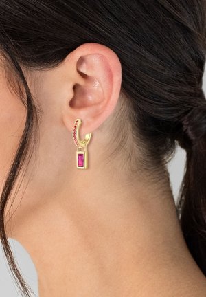 Goldene Ohrringe mit einem geschwungenen Design und rosa Edelsteinen, darunter ein rechteckiger, rubinähnlicher Stein, hängen neben dem Ohr.