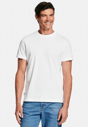 Man smiling, wearing a plain white short-sleeve crew neck t-shirt and blue jeans, standing against a white background.