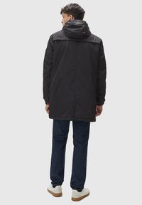 Svart, lång vinterjacka med huva. Har en mjuk yta, justerbara ärmar och en ventil bak. Bärs över blå jeans och vita sneakers.