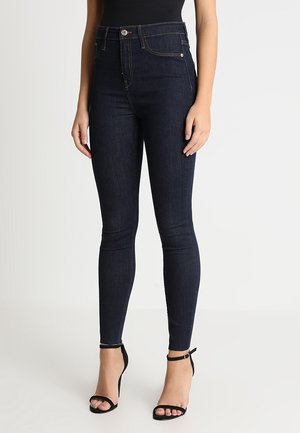 Slim-fit dark blue jeans worn by a person standing, paired with black ankle-strap high-heeled sandals and a black top.