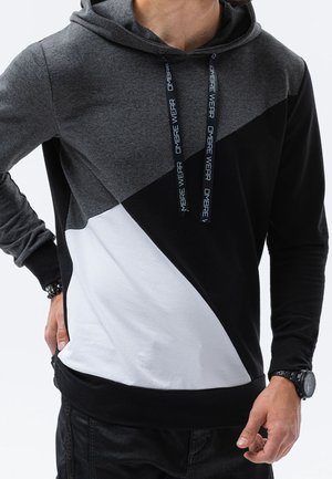 Man draagt een color-block hoodie met donkergrijze, zwarte en witte diagonale secties en zwarte trekkoorden met de tekst "OMBRE WEAR".