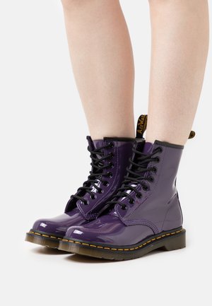 Bottes montantes à lacets en cuir verni violet avec lacets noirs, couture jaune sur la semelle et une languette de traction marquée à l'arrière.