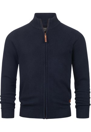 Marineblå strikket cardigan med høy krage, hel glidelås, ribbestrikkede mansjetter og kant, med et lærmerke nær kanten.