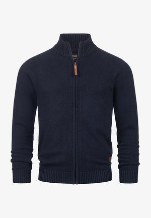 Sininen neulecardigan, jossa on korkea kaulus, täysipitkä vetoketju, ribbireunat hihoissa ja helmassa, sekä nahkainen tagi helman lähellä.