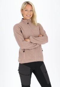 Jasnoróżowa polarowa kurtka z wysokim kołnierzem, zapinanymi kieszeniami i teksturowaną powierzchnią. Noszona z czarnymi wielofunkcyjnymi spodniami z zamkami.