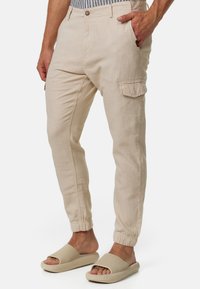 Pantalones cargo de lino beige claro con puños elásticos, bolsillos laterales y cierre de botón. Combinados con sandalias beige de punta abierta.