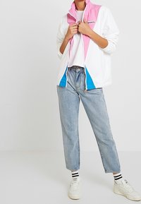 Biała lekka kurtka z różowymi i niebieskimi akcentami, z wysokim kołnierzem, zamek błyskawiczny oraz luźny krój, połączona z jasnoniebieskimi dżinsami.