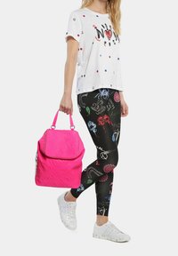 Femme portant un T-shirt blanc et des leggings noirs à motifs, tenant un sac à main rose vif à poignées courtes, marchant en baskets blanches.