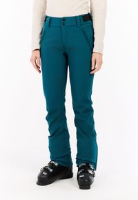 Žalios slidinėjimo kelnės, pagamintos iš tamprios medžiagos, su siaurėjimu, užtrauktukais kišenėse ir reguliuojamu juosmeniu, kuriame yra du mygtukai. Apsirengtos su juodais slidinėjimo batais.