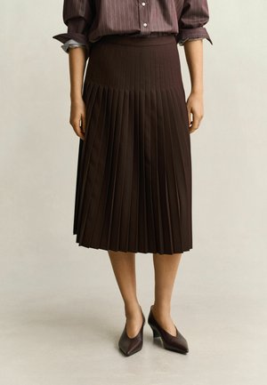 Femme portant une jupe midi plissée marron foncé, des talons pointus assortis et une chemise rayée à manches longues retroussées.