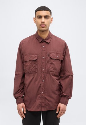 Giovane uomo con capelli corti che indossa una camicia bordeaux con bottoni, due tasche sul petto e una tasca con cerniera, su uno sfondo neutro.