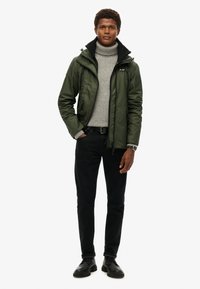 Grüne Zip-Jacke mit schwarzem Kragen über einem hellgrauen Rollkragenpullover, kombiniert mit schwarzen Jeans und schwarzen Schuhen. Schlichtes Design, keine Muster.