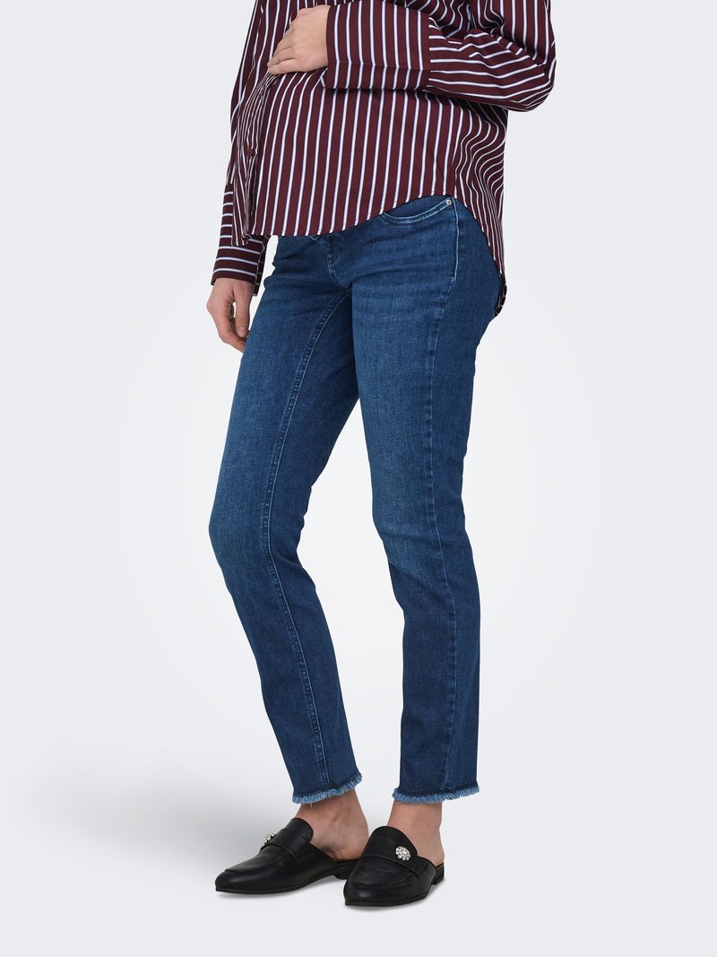 Personne portant un jean bleu foncé à ourlet effiloché, des mocassins noirs sans lacets avec des ornements, et une chemise à manches longues rayée bordeaux et blanche.