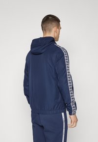 Giacca con cappuccio blu navy con polsini e cintura elasticizzati, dotata di strisce laterali geometriche bianche e una texture liscia.