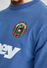 Sudadera de algodón azul con un gran logo blanco de la marca y un parche bordado circular con "Obey Societa" y una figura negra rodeada de ramas de olivo.