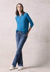 Türkisblaue Bluse mit langen Ärmeln und Kragen, kombiniert mit blauen Schlagjeans und weißen Sneakern, vor einem neutralen Hintergrund.