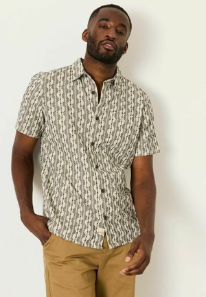 Mann mit Bart, der ein kurzärmeliges gemustertes Hemd mit Knöpfen und beige Hose trägt, steht vor einem einfarbigen hellen Hintergrund.