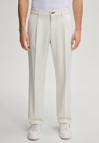 Cremefarbene Hosen mit lockerer Passform, ausgestattet mit einem gefalteten Vorderteil, Gürtelschlaufen und einem Knopfverschluss. Tragen mit weißen Sneakers.