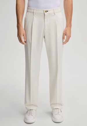 Cremefarbene Hosen mit lockerer Passform, ausgestattet mit einem gefalteten Vorderteil, Gürtelschlaufen und einem Knopfverschluss. Tragen mit weißen Sneakers.