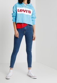 Felpa cropped azzurro chiaro con logo "Levi's" in bianco e rosso, indossata sopra una maglietta rossa. Jeans attillati blu scuro, abbinati a sneaker bianche.