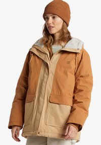 Brun och beige isolerad jacka med stora fickor, en krage med fuskpäls och dragkedja. Matchad med en brun stickad mössa.