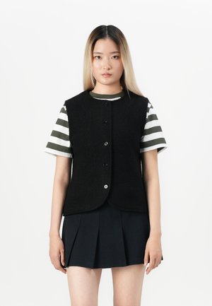 Jeune femme portant un gilet noir à boutons sur un t-shirt rayé vert et blanc et une jupe plissée noire, se tenant devant un fond blanc.