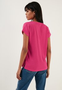 Pink textured short-sleeve top with side slits, paired with blue denim jeans. The back features a rounded neckline and relaxed fit.