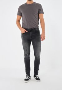 Hombre con una camiseta gris ajustada, jeans skinny negros desgastados y zapatillas en blanco y negro de pie con una mano en el bolsillo.
