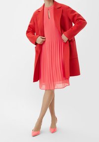 Manteau en laine rouge par-dessus une robe plissée rose avec un col en trou de serrure. Le mannequin porte des talons aiguilles roses vifs. Tissu lisse et silhouette structurée.