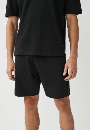 Homme portant un t-shirt noir uni à manches courtes et un short noir, debout sur un fond neutre.