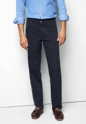 Mann, der dunkelblaue Hose, hellblaues langärmeliges Hemd mit hochgekrempelten Ärmeln und braune Lederschuhe trägt, steht vor weißem Hintergrund.
