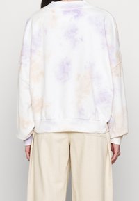 Person som bär en lös vit sweatshirt med ljuslila och beige batikmönster, kombinerad med högmidjade beige byxor.