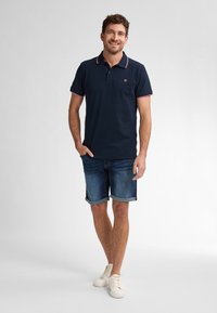 Camiseta polo azul marino con acentos rojos, combinada con shorts de mezclilla. El modelo está de pie con las manos en los bolsillos, usando zapatillas blancas.