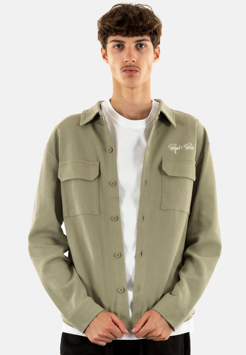 Camisa de corte grande en verde oliva con dos bolsillos en el pecho, cierre de botones y textura acanalada. Llevada sobre una camiseta blanca.