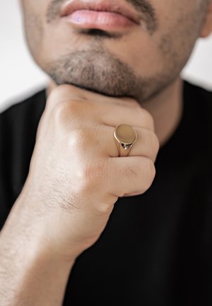 Gold-colored ring with a smooth, round top, resting on a hand with a natural skin texture. The person is wearing a black shirt.