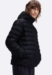 Giacca a vento nera con cappuccio, con sezioni trapuntate, zip frontale e un piccolo logo. Il materiale sembra liscio e isolato.